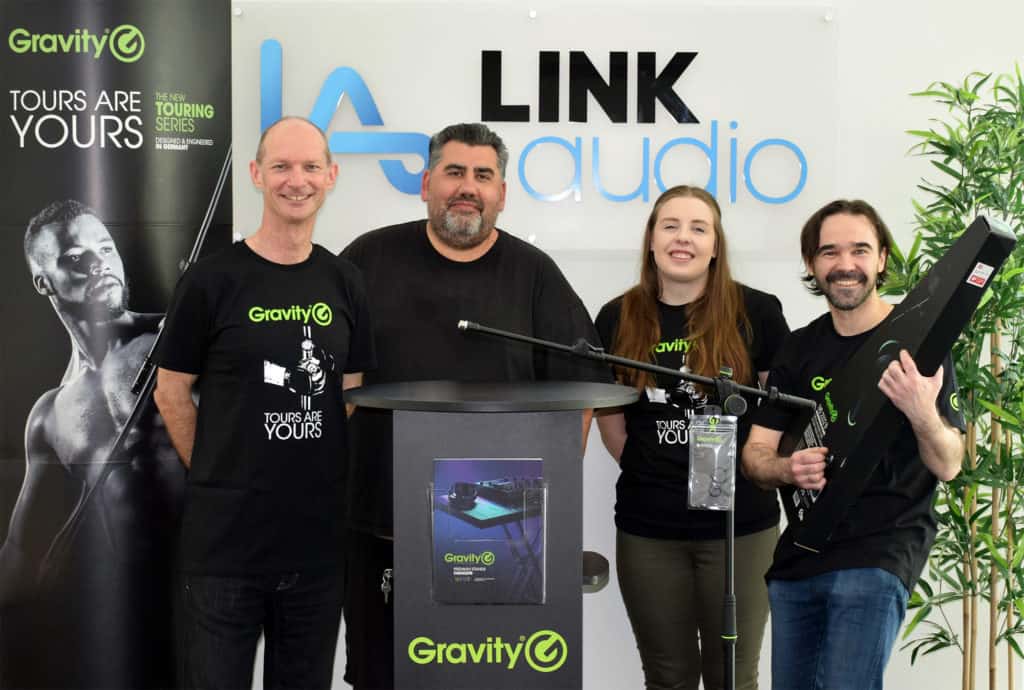 AHG-Gravity_LinkAudio Vier Personen stehen in einem Innenraum hinter einem Podium mit dem Gravity-Logo und lächeln in die Kamera, während im Hintergrund Veranstaltungstechnik und Werbebanner zu sehen sind.