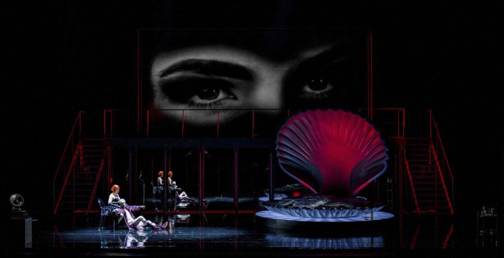 KLEIN2ETC-Series3-PowderHerFace-21 Eine Bühnenszene mit Darstellern in dramatischen Kostümen, einem großen roten, muschelförmigen Bett und einem Schwarz-Weiß-Bild von Augen, das über das Bühnenbild projiziert wird.