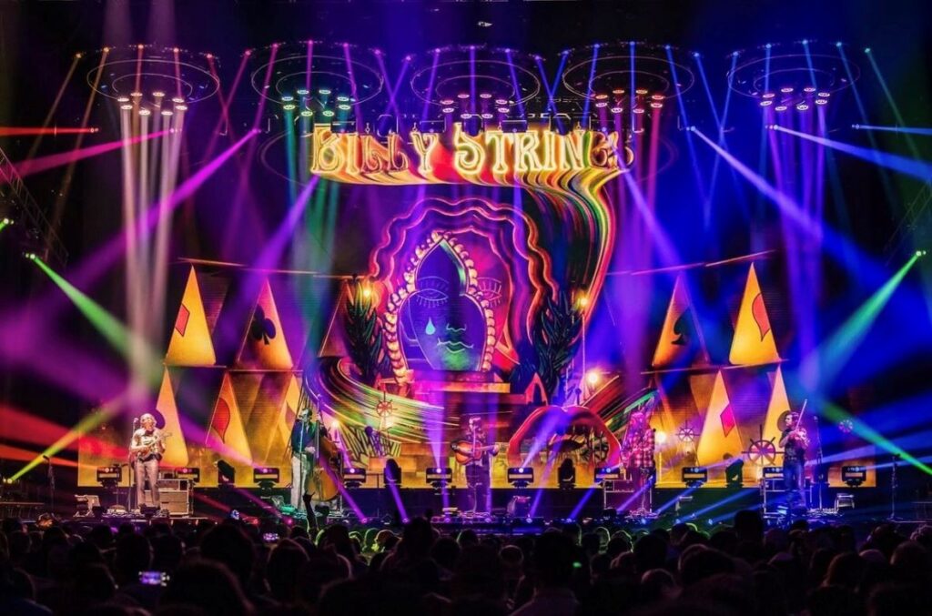 Billy Strings 3 ┬® Jesse Faatz Eine Band spielt auf der Bühne mit leuchtenden, bunten Lichtern und einer psychedelischen Kulisse, die Billy Strings zeigt. Im Vordergrund ist das Publikum zu sehen.
