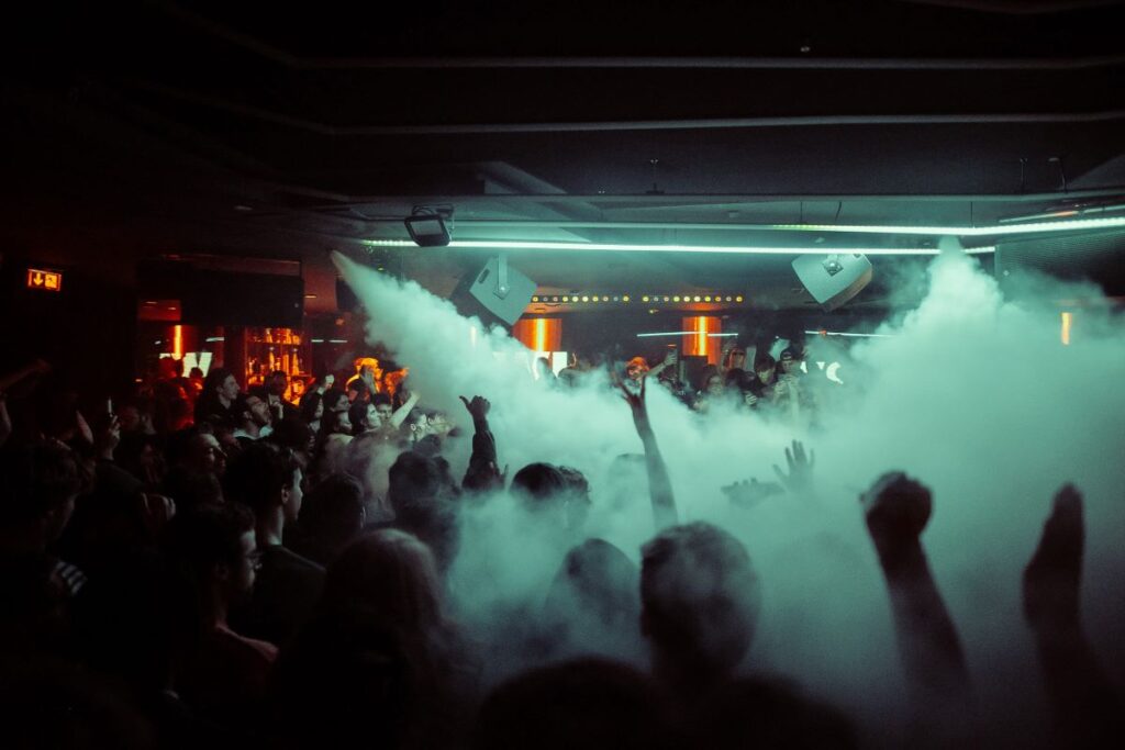 CAD_Pacha2 Ein überfüllter Nachtclub mit Menschen, die ihre Hände heben, während Rauch in der Luft liegt, beleuchtet von schummrigen, bunten Lichtern und Neonakzenten.