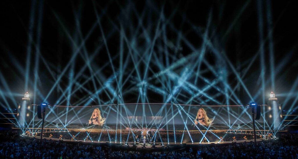 CopyrightSTUFISH_1312-AA-Munich_02AUG24_7400 sml Eine große Konzertbühne mit hellen Scheinwerfern, eine Menschenmenge im Publikum und das Bild eines Sängers, das nachts auf zwei große Leinwände über der Bühne projiziert wird.