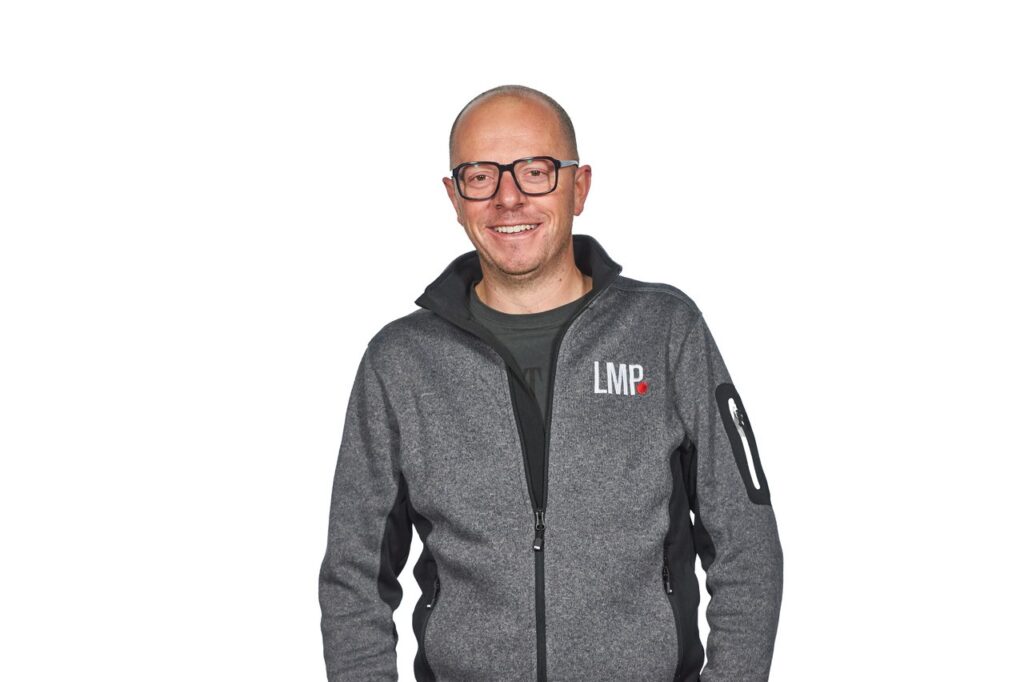 Eine Person mit Brille und einer grauen Jacke mit Reißverschluss und der Aufschrift LMP steht vor einem schlichten weißen Hintergrund und lächelt in die Kamera.