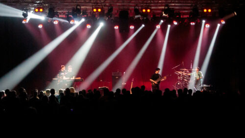 Eine Band tritt auf der Bühne unter hellen Scheinwerfern auf. Professionelle Veranstaltungstechnik verstärkt die Show, während das Publikum zwischen Musikinstrumenten und Equipment zuschaut.