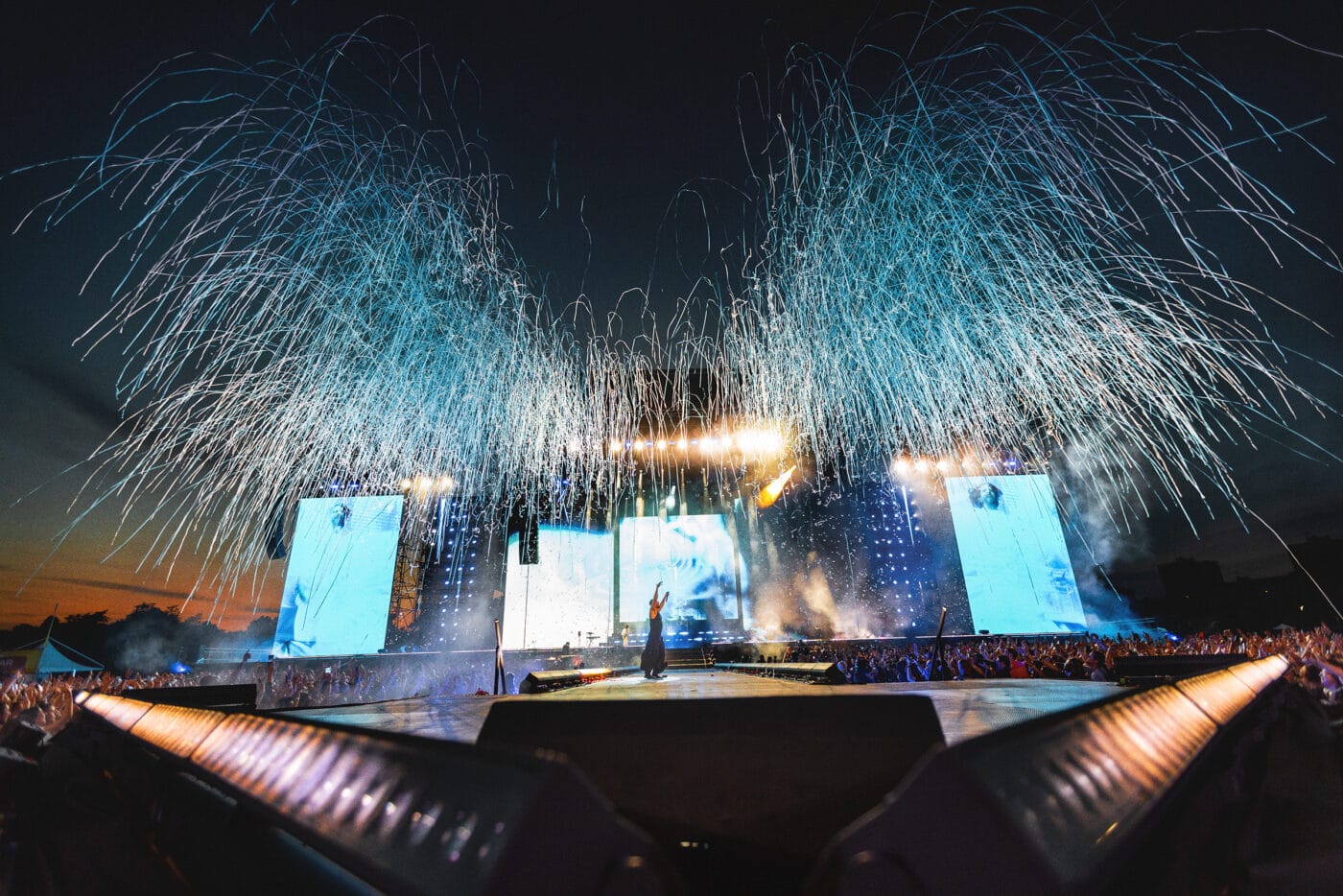 250527_ImagineDragons_LoomTour_Milan_1778_copy Ein Musiker tritt nachts auf einer großen Freilichtbühne auf, mit blauem und weißem Feuerwerk und Lichtern, und eine Menschenmenge schaut zu.