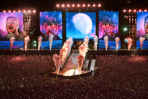 Eine große Menschenmenge besucht ein nächtliches Konzert mit Raucheffekten, großen Bildschirmen und einem Künstler, der auf einer großen Bühne steht.