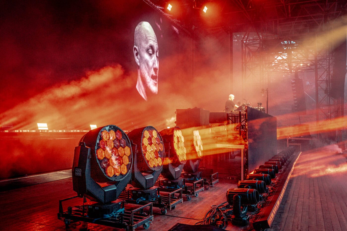 2025 paul kalkbrenner location: wuhlheide Ein Künstler steht an einem DJ-Pult auf der Bühne mit orangefarbenen Lichtern und Nebel, verstärkt durch Veranstaltungstechnik, während ein großes projiziertes Gesicht auf der Leinwand hinter ihm erscheint.