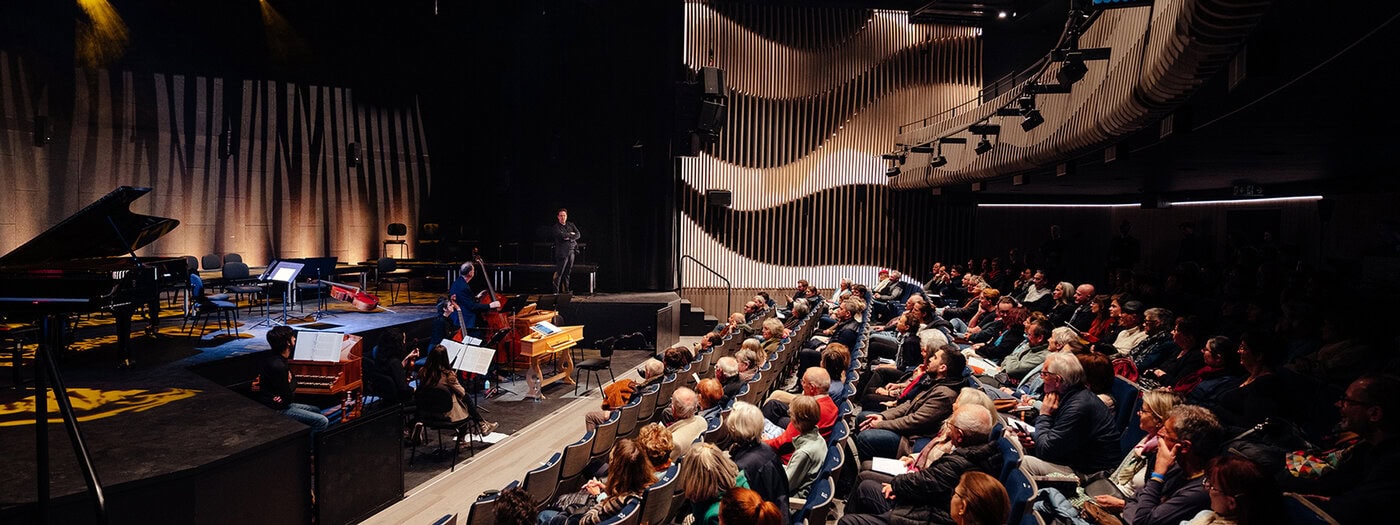 Ein dicht gedrängtes Publikum sitzt in einem modernen Konzertsaal und sieht zu, wie Musiker auf der Bühne mit verschiedenen Instrumenten unter Bühnenbeleuchtung spielen.