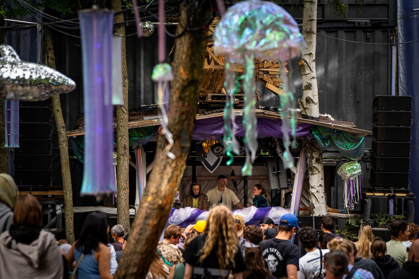 Eine Menschenmenge beobachtet eine Gruppe von Menschen auf einer kleinen Freiluftbühne, die mit lila Stoff und Quallenornamenten geschmückt ist, umgeben von Bäumen und hängenden Dekorationen.