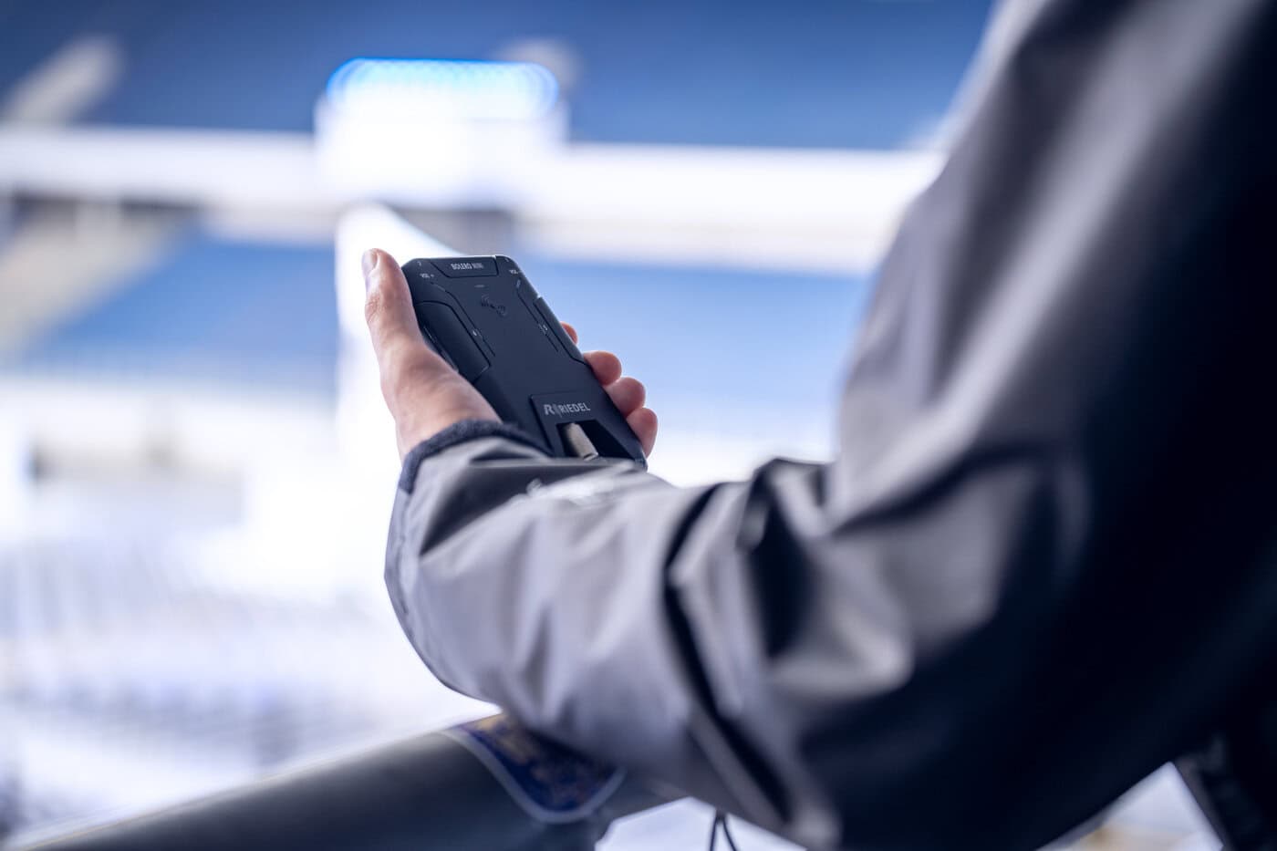 Eine Person in einer grauen Jacke hält ein schwarzes, robustes Smartphone im Freien in der Nähe eines Metallgeländers, mit verschwommenen Stadionsitzen im Hintergrund.