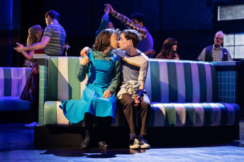 Zwei Menschen sitzen eng beieinander und küssen sich an einem Retro-Diner-Stand, während andere Menschen im Hintergrund bei Bühnenbeleuchtung reden und tanzen.