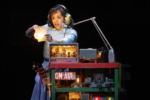 Eine Person mit Kopfhörern spricht in ein Mikrofon, während sie eine Glühbirne und Papier in der Hand hält. Sie sitzt an einem Schreibtisch mit alten elektronischen Geräten und einem ON AIR-Schild.