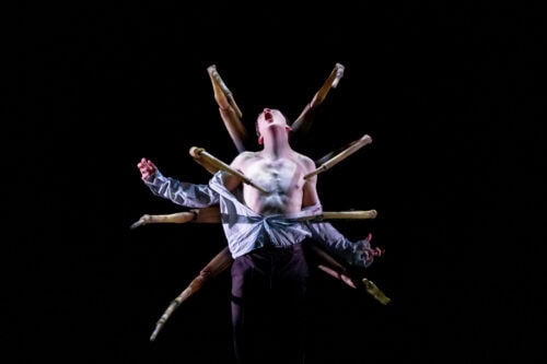 Eine hemdsärmelige Person steht mit offenem Mund, umgeben von mehreren ausgestreckten Armen, vor einem schwarzen Hintergrund und erzeugt einen dramatischen und surrealen visuellen Effekt.