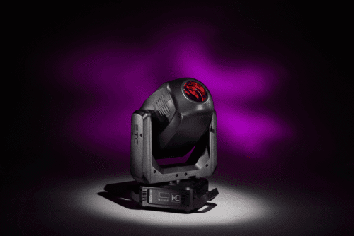 Ein schwarzer Bühnenscheinwerfer mit einer roten Linse projiziert ein violettes Lichtmuster auf einen dunklen Hintergrund. Das Gerät steht auf einer ebenen Fläche und ist leicht nach oben gerichtet.