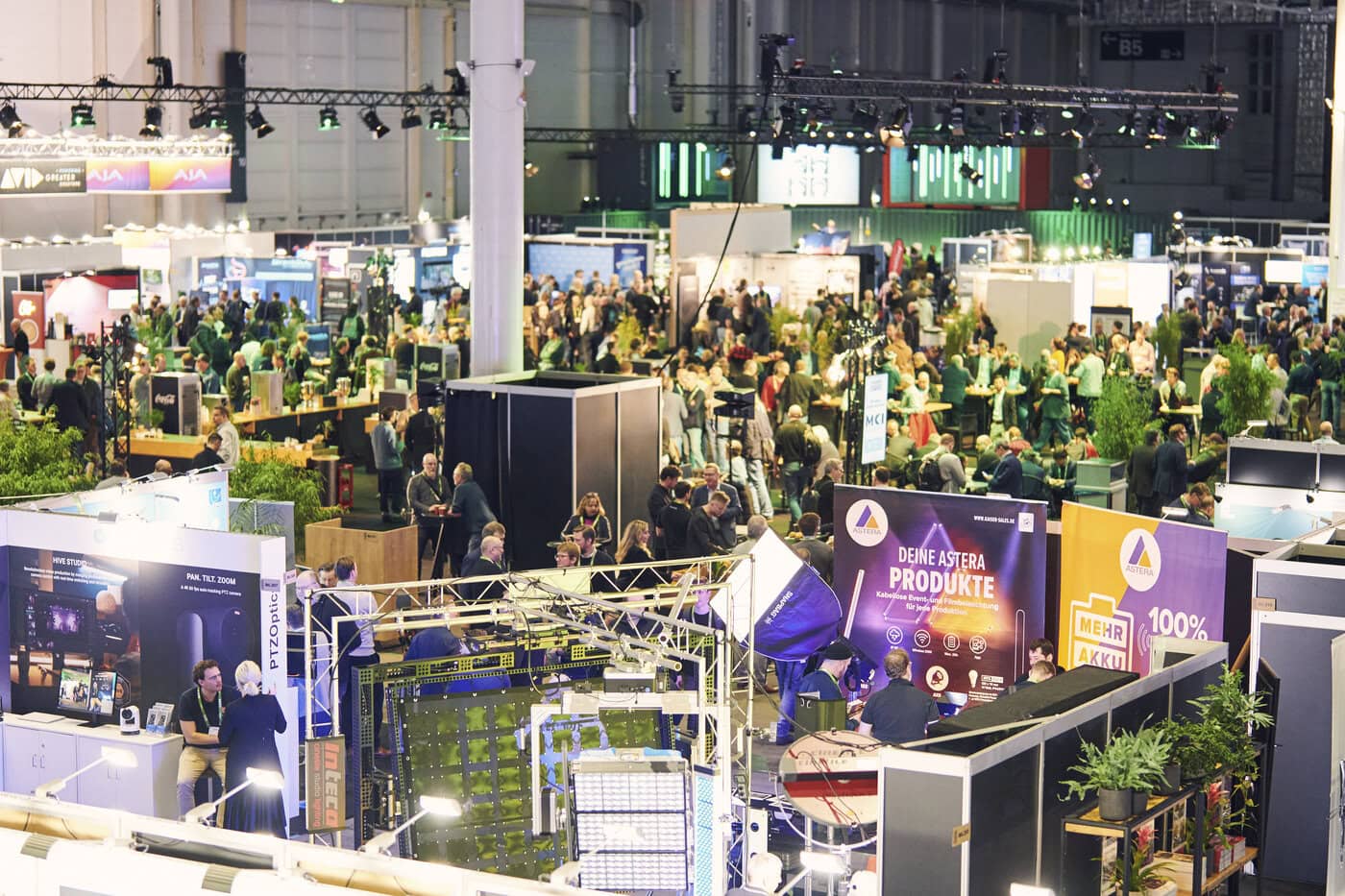 Eine große überdachte Messe oder ein Kongress mit vielen Menschen, die verschiedene Stände besuchen, die mit Bannern, Schildern und Grünpflanzen geschmückt sind.