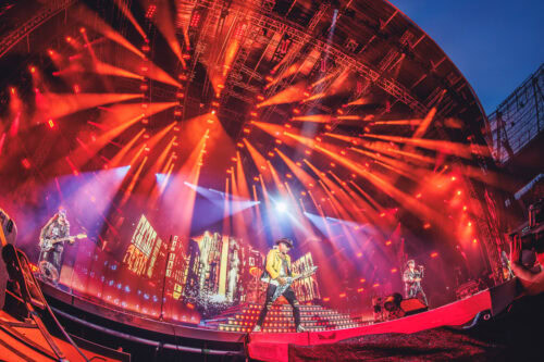 Eine Rockband tritt auf einer großen, hell erleuchteten Bühne mit orangefarbenen Scheinwerfern und einer Stadtkulisse auf, die aus einem niedrigen Winkel betrachtet wird.