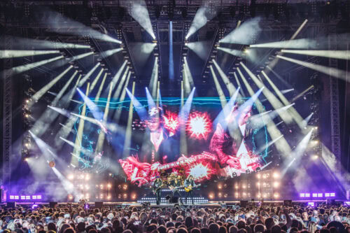 Eine große Menschenmenge besucht ein Konzert mit einer hell erleuchteten Bühne, farbenfrohen Lichtern und Bildschirmen, auf denen Grafiken und visuelle Darstellungen hinter einer auftretenden Band zu sehen sind.