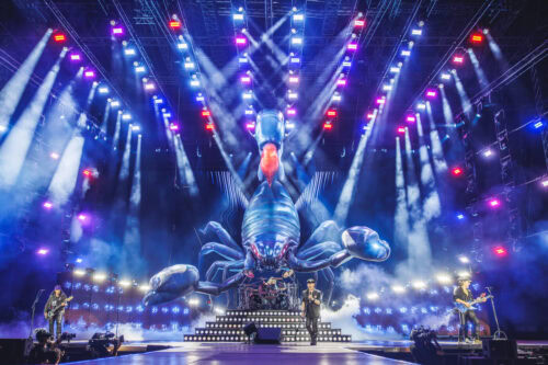 Eine Rockband spielt auf der Bühne mit bunten Lichtern und einer großen, detaillierten Skorpion-Bühnenrequisite hinter ihnen, umgeben von Nebel- und Raucheffekten.