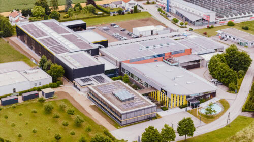 Luftaufnahme eines großen Industrie- oder Gewerbekomplexes mit mehreren Gebäuden, Sonnenkollektoren auf den Dächern, Parkplätzen und umliegenden Grünflächen.