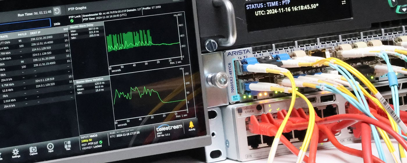 startbanner_PMIP_2024 Eine Nahaufnahme von Netzwerkgeräten mit angeschlossenen bunten Ethernet-Kabeln neben einem Monitor, der Datengrafiken und Informationen zum Systemstatus anzeigt.