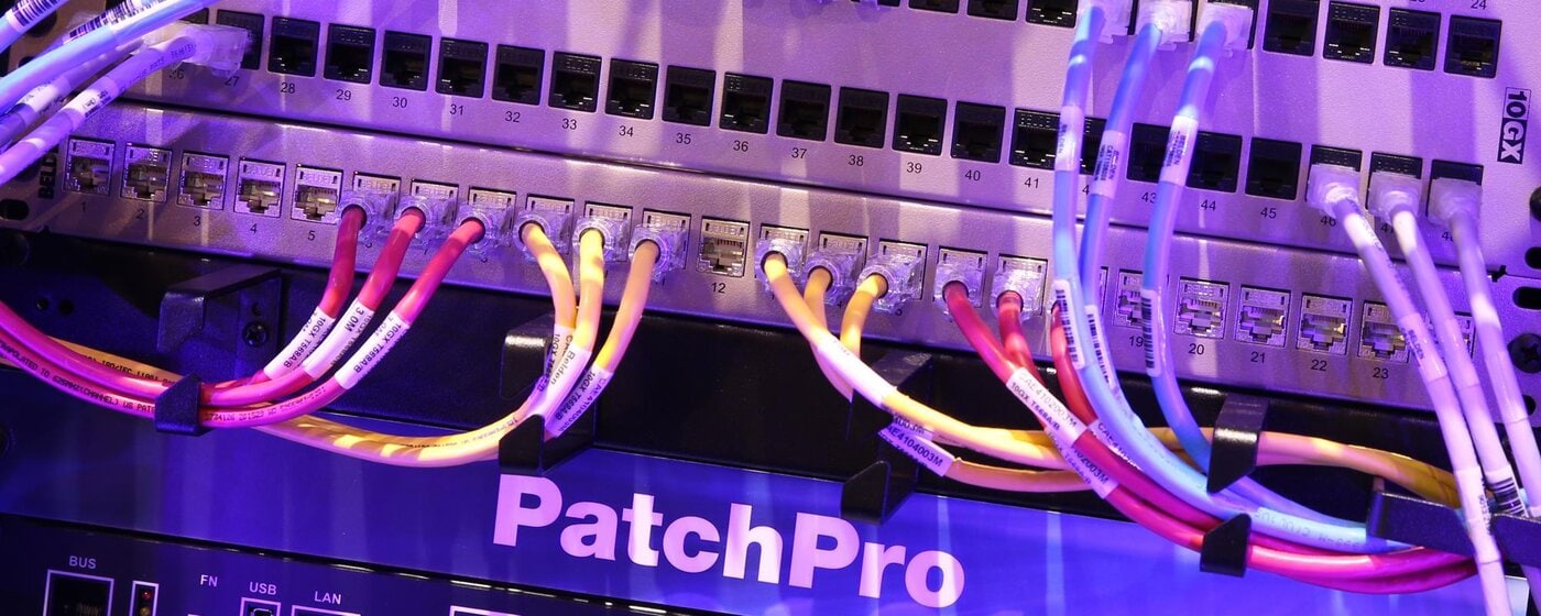 startbanner_netzwerkpraxis Eine Nahaufnahme eines PatchPro-Netzwerk-Switchpanels mit mehrfarbigen Kabeln, die an nummerierten Anschlüssen unter blauer und violetter Beleuchtung angeschlossen sind.