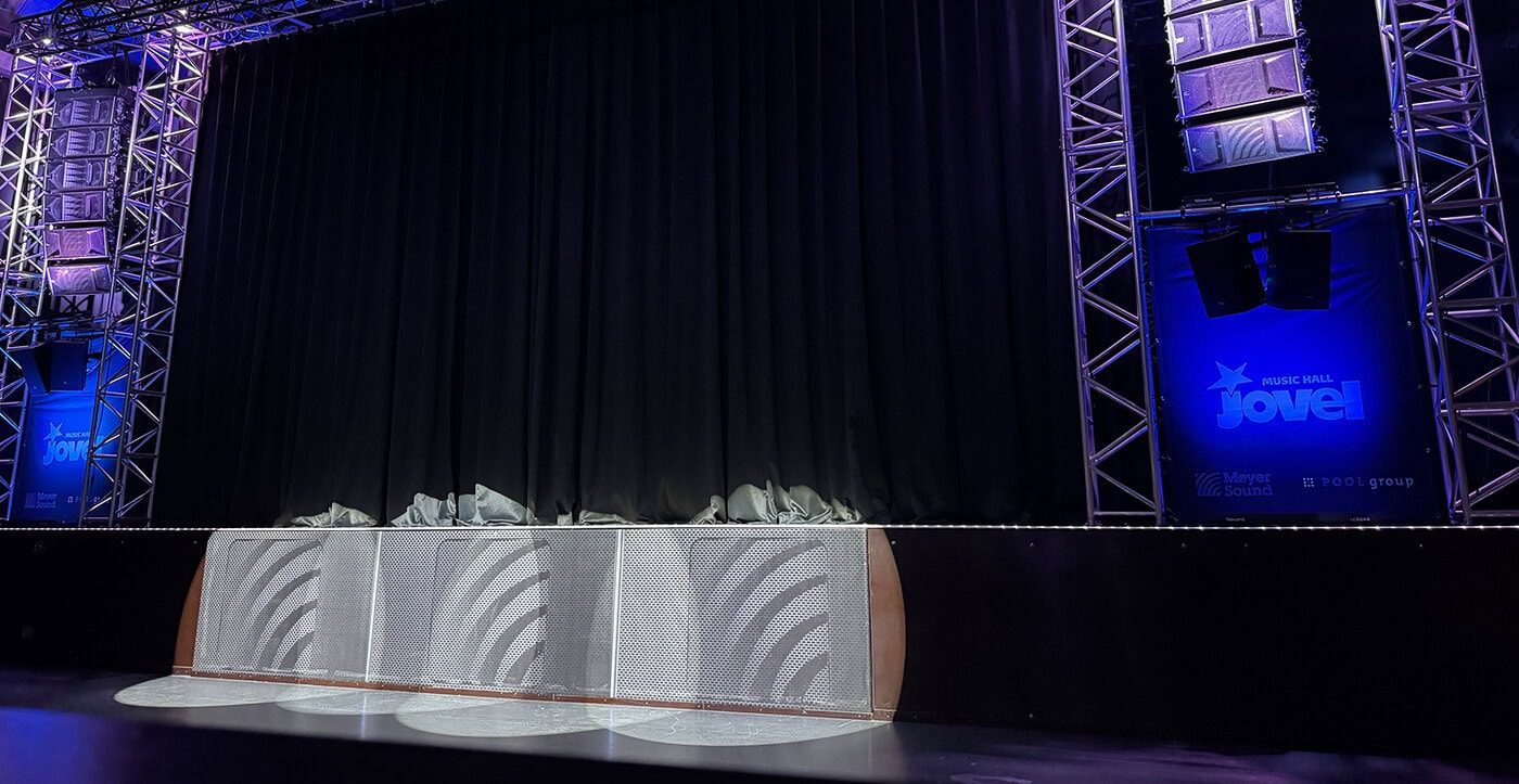 Eine Bühne mit einem geschlossenen schwarzen Vorhang, umgeben von einem Metallgerüst und blauer Beleuchtung, mit Scheinwerfern, die auf den Boden davor scheinen.