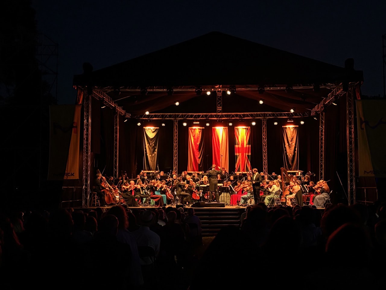 AD_DtPhilharmonieMerck3 Ein Sinfonieorchester spielt auf einer nächtlichen Freilichtbühne, die Musiker sitzen im Scheinwerferlicht, und an der Rückseite der Bühne hängen Fahnen. Das Publikum ist im Schatten sichtbar.