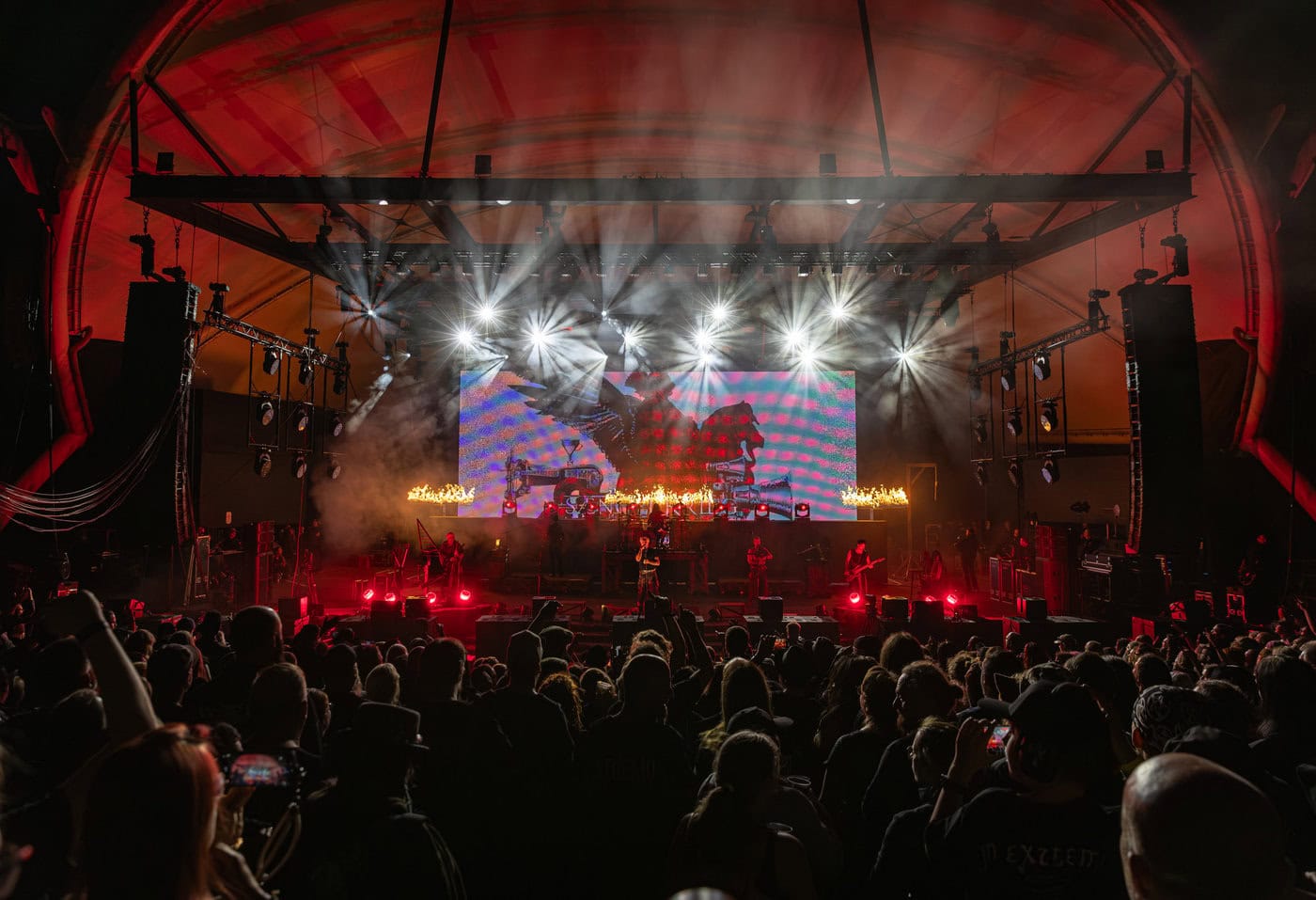 Ein Publikum verfolgt ein Live-Konzert an einem Veranstaltungsort im Freien mit hellen Bühnenlichtern, Raucheffekten und einer großen Leinwand, auf der Bilder hinter der Band angezeigt werden.