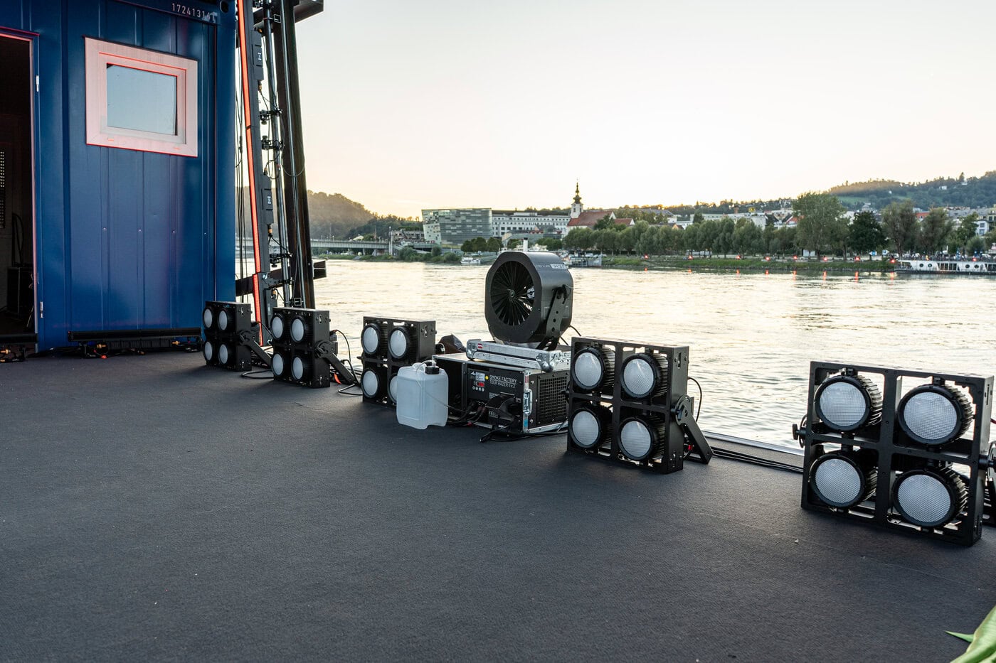 Bühnenbeleuchtung und ein großer Ventilator auf einer Bühne an einem Fluss, mit Bäumen und Gebäuden am gegenüberliegenden Ufer im Hintergrund.