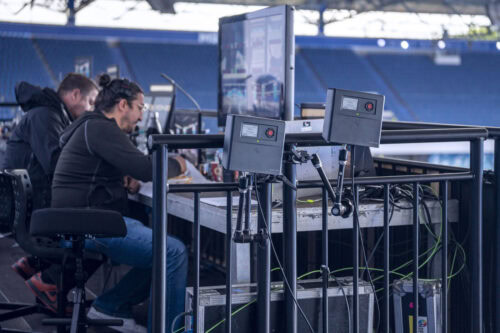 Zwei Personen sitzen an einem Tisch mit Monitoren und elektronischen Geräten in einer Stadionumgebung, wobei Kabel und Geräte im Vordergrund zu sehen sind.