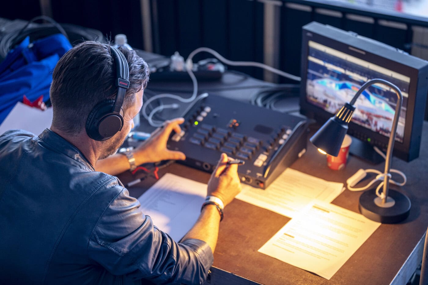 Eine Person mit Kopfhörern bedient ein Audio- oder Videogerät an einem Schreibtisch mit Papieren, einem Monitor, der eine Live-Übertragung zeigt, und einer Schreibtischlampe.