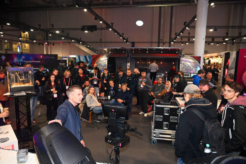 Eine Gruppe von Menschen sitzt und steht um einen Stand bei einer belebten Indoor-Veranstaltung und beobachtet eine Vorführung mit elektronischen Geräten und Bildschirmen.