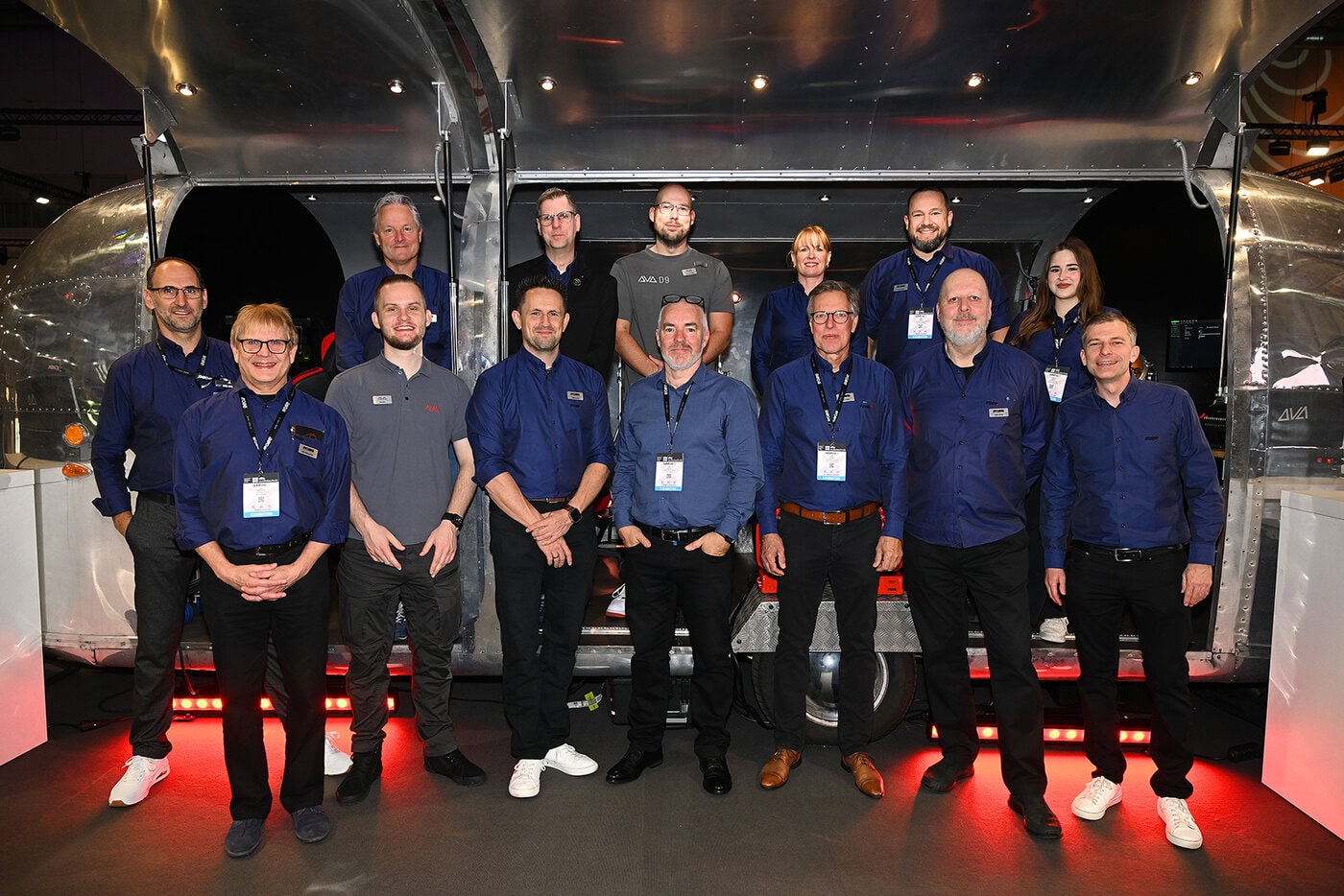 Vierzehn Personen stehen gemeinsam in einem Innenraum vor einem glänzenden Metallanhänger, tragen Business Casual oder Markenhemden und lächeln für ein Gruppenfoto.