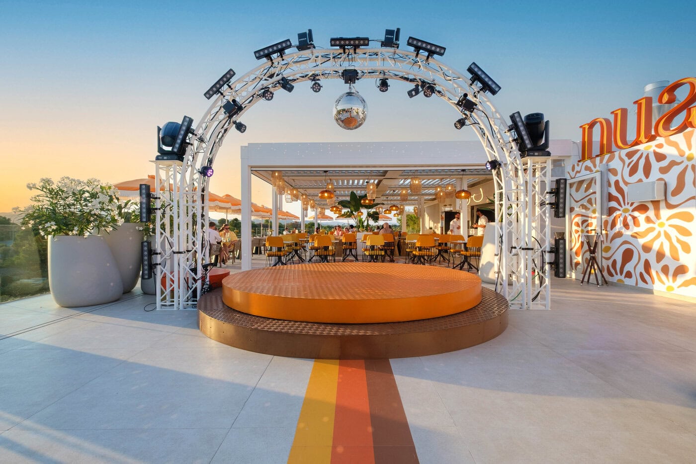 Ein Außenrestaurant mit orange-weißem Dekor, einer runden Bühne unter einem Metallbogen mit Lichtern, Stühlen, Tischen und Topfpflanzen im Hintergrund.