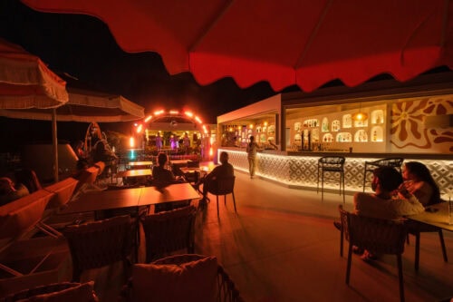 Bar und Lounge im Freien bei Nacht, mit Tischen und einem gut beleuchteten Bartresen; im Hintergrund ist eine Bühne mit Lichtern und Künstlern zu sehen.
