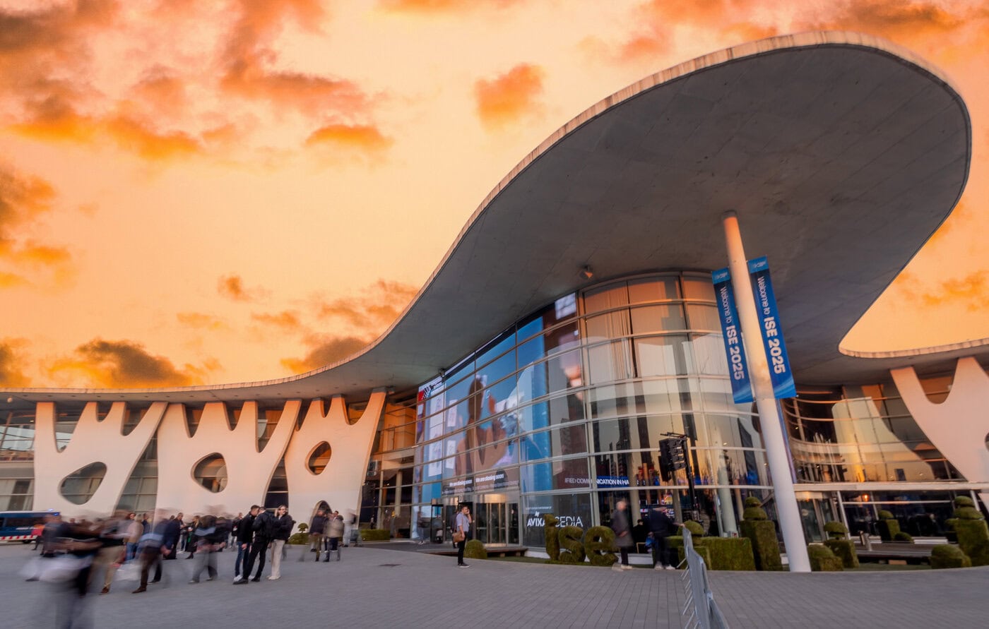 Modernes Gebäude mit geschwungenem Dach und großen Glasfenstern unter einem orangefarbenen Himmel, mit Menschen, die vor dem Eingang gehen und stehen.