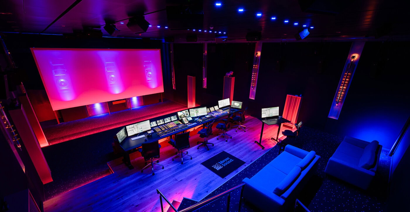Ein moderner Regieraum mit mehreren Monitoren und Tonmischgeräten steht vor einer großen Leinwand; blaue und rosafarbene Beleuchtung erhellt den Raum mit Sitzgelegenheiten im Vordergrund.