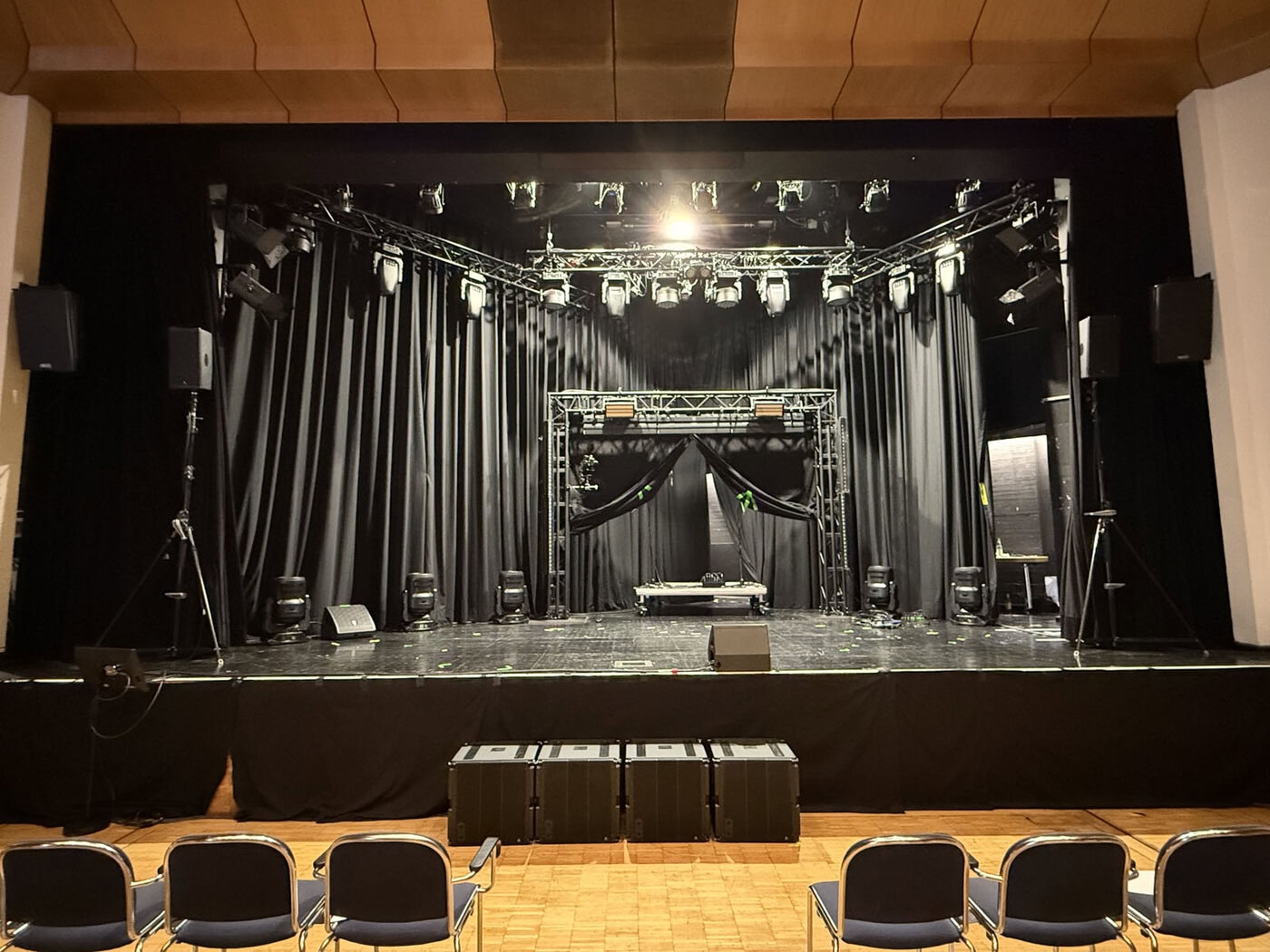 Blick auf eine leere Theaterbühne mit schwarzen Vorhängen, Bühnenbeleuchtung und Tontechnik; im Vordergrund sind mehrere leere Stühle zu sehen.