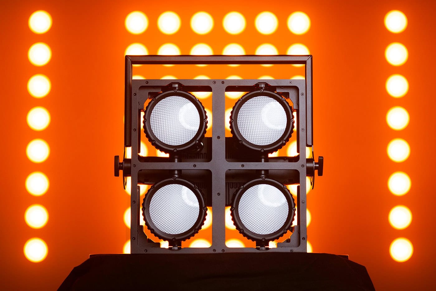 Eine quadratische Anordnung von vier Bühnenscheinwerfern ist vor einem Gitter aus leuchtend orangefarbenen Lichtern positioniert, die sich alle vor einem orangefarbenen Hintergrund befinden.