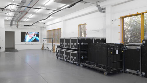 Ein geräumiger, weißer Raum mit Gerätekoffern auf Rädern, die entlang einer Wand gestapelt sind, und einem großen abstrakten Bild auf einem Monitor am anderen Ende.