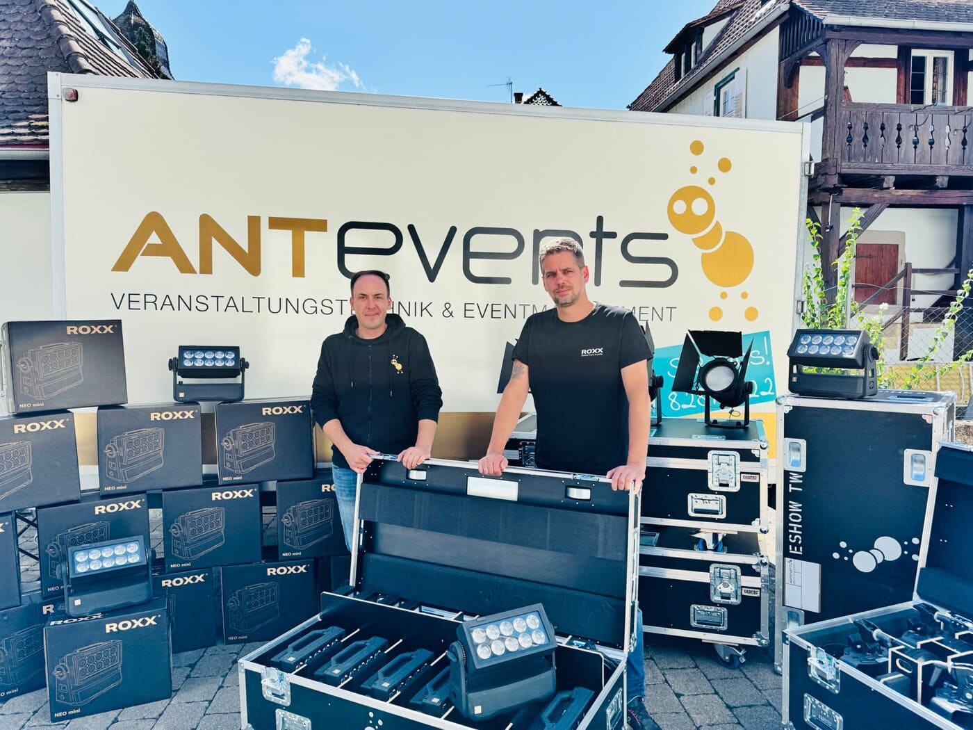 Zwei Personen stehen vor einem weißen ANTevents-Truck, umgeben von Beleuchtungsausrüstung und Koffern, mit Gebäuden im Hintergrund.