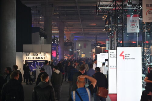 Eine überfüllte Messehalle mit Menschen, die zwischen Ständen und Schildern umherlaufen, darunter solche mit der Aufschrift Behind the Excitement und Area Four Industries.
