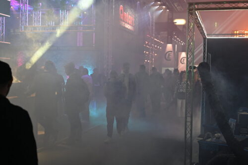Die Menschen gehen durch einen nebligen, farbenfrohen Veranstaltungsraum mit hellen Lichtern, Metalltraversen und verschiedenen Ständen im Hintergrund.