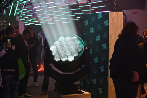 Ein großes Bühnenlicht sendet schräge Strahlen aus und beleuchtet einen Stand mit Mad Max-Branding. Die Menschen stehen und interagieren um das Display herum in einem überdachten Veranstaltungsraum.