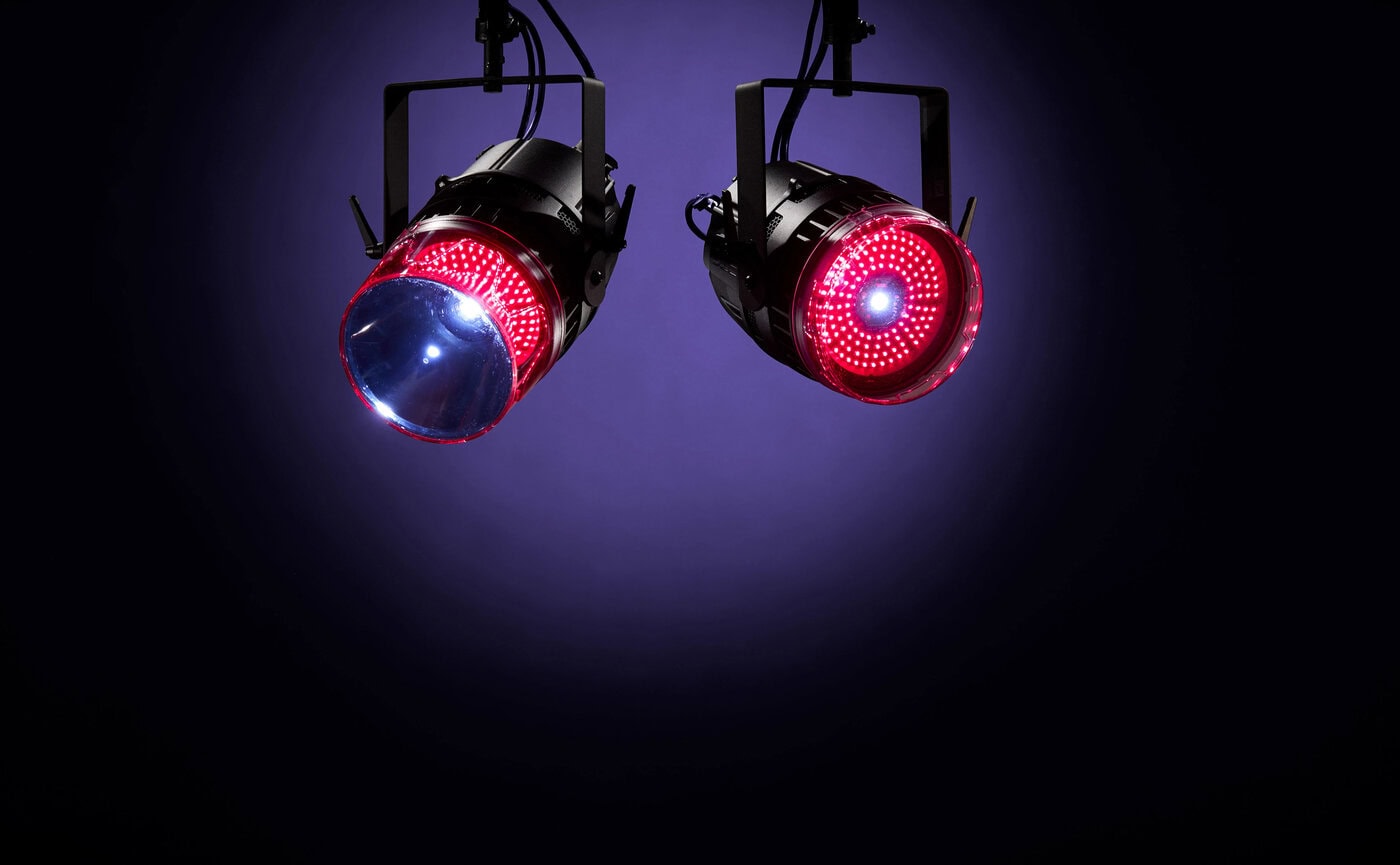 Zwei schwarze Bühnenscheinwerfer, die von der Decke hängen und jeweils rotes Licht ausstrahlen, vor einem dunkelvioletten Hintergrund.