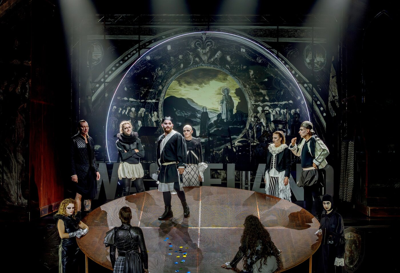 Eine Gruppe von Schauspielern in historischen Kostümen steht auf einer runden Bühne, die von einem dramatischen gemalten Hintergrund und theatralischer Beleuchtung untermalt wird.