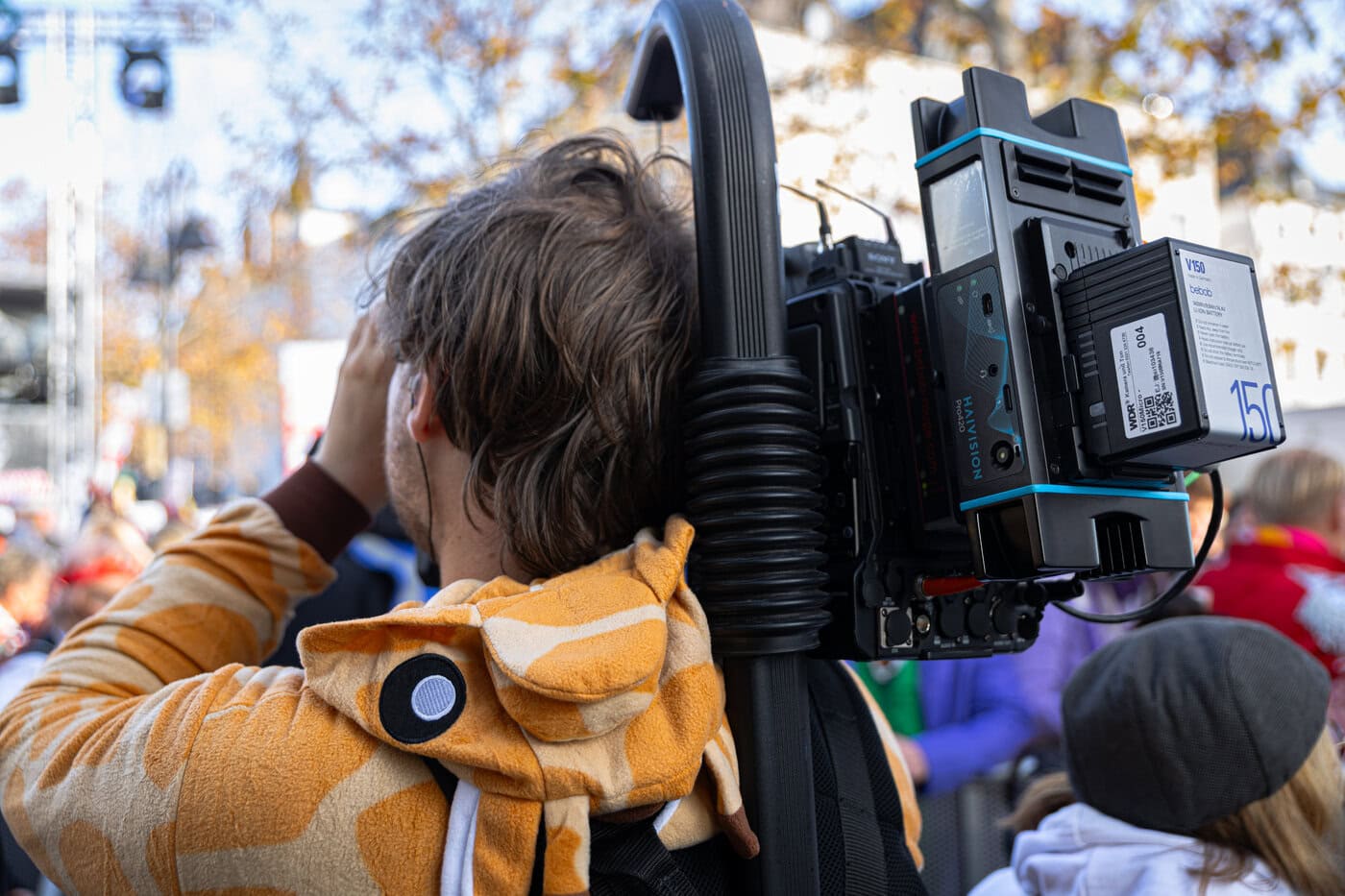 Eine Person in einem Strampelanzug mit Giraffenmuster bedient eine professionelle Kamera mit einem großen Akkupack bei einer Veranstaltung im Freien mit einer Menschenmenge im Hintergrund.