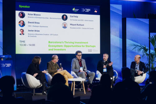 Vier Männer sitzen auf einem Podium und diskutieren unter einem Bildschirm, auf dem ihre Namen, Titel und das Thema angezeigt werden: Barcelonas florierendes Investitions-Ökosystem.