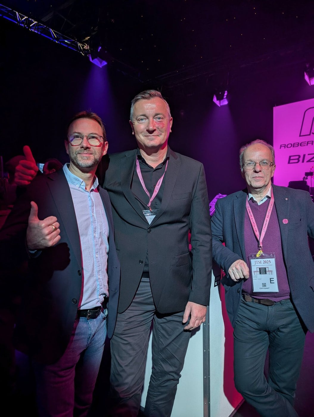 Drei Männer in Anzügen und mit Schlüsselbändern für Veranstaltungen stehen in einem Innenraum unter violettem Licht zusammen, wobei einer von ihnen den Daumen nach oben streckt.