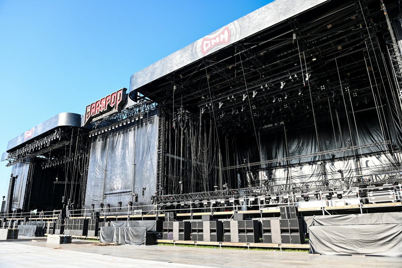 Eine große Konzertbühne im Freien mit schwarzen Vorhängen, Beleuchtung, Lautsprechern und einem Graspop-Schild, fotografiert unter einem klaren blauen Himmel.