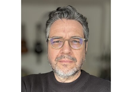 SpeakerImage_Mirko Herth_Original.1a410bfe Ein Mann mittleren Alters mit grauem Haar, Brille und Bart trägt einen dunklen Pullover und schaut direkt in die Kamera. Der Hintergrund ist unscharf.